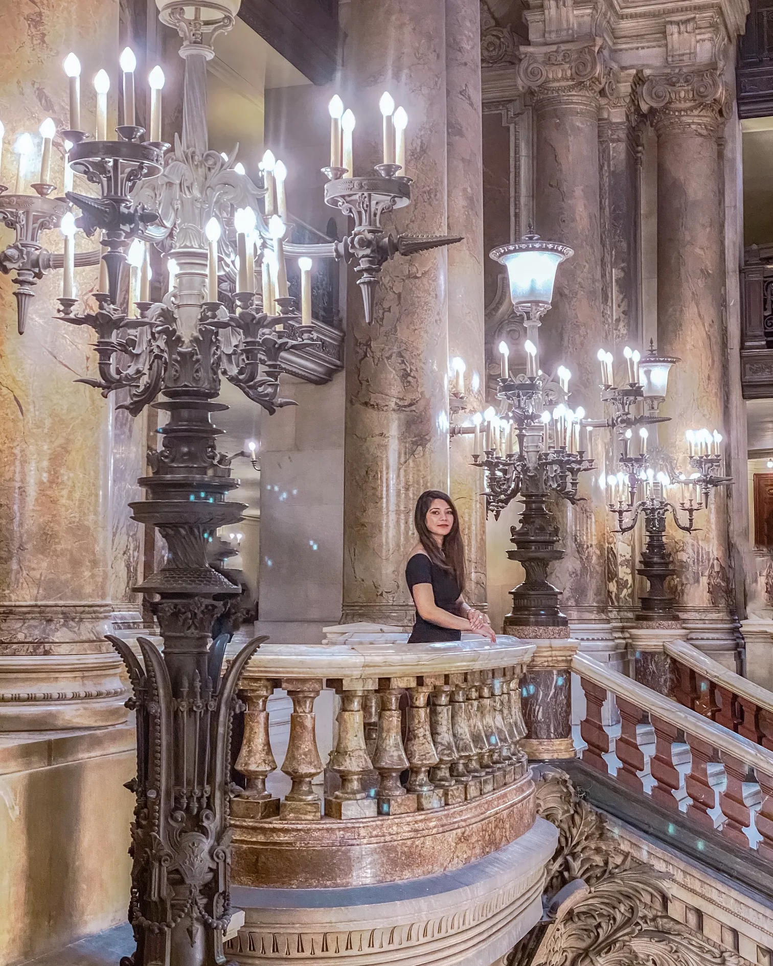 Woman in a black dress standing on an ornate marble balcony inside a grand, candlelit historic theatre or opera house.