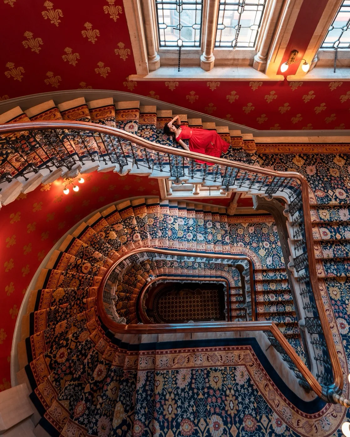Woman in a red dress reclining on an ornate carpeted spiral staircase beneath tall stained-glass windows.