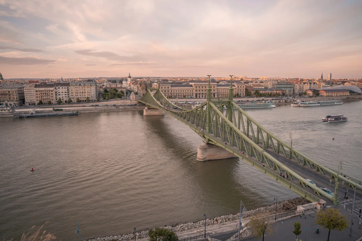 Green Liberty Bridge spanning the Danube at sunset with city buildings and riverboats in the background.