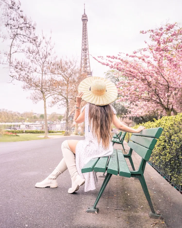 Woman in a summer dress and wide straw hat sitting on a green park bench facing the Eiffel Tower amid pink blossoms.