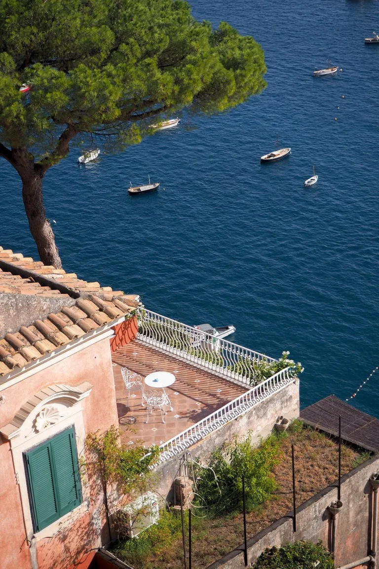 Terraced Mediterranean villa with a small wrought-iron table and chairs overlooking a deep-blue sea dotted with small boats.