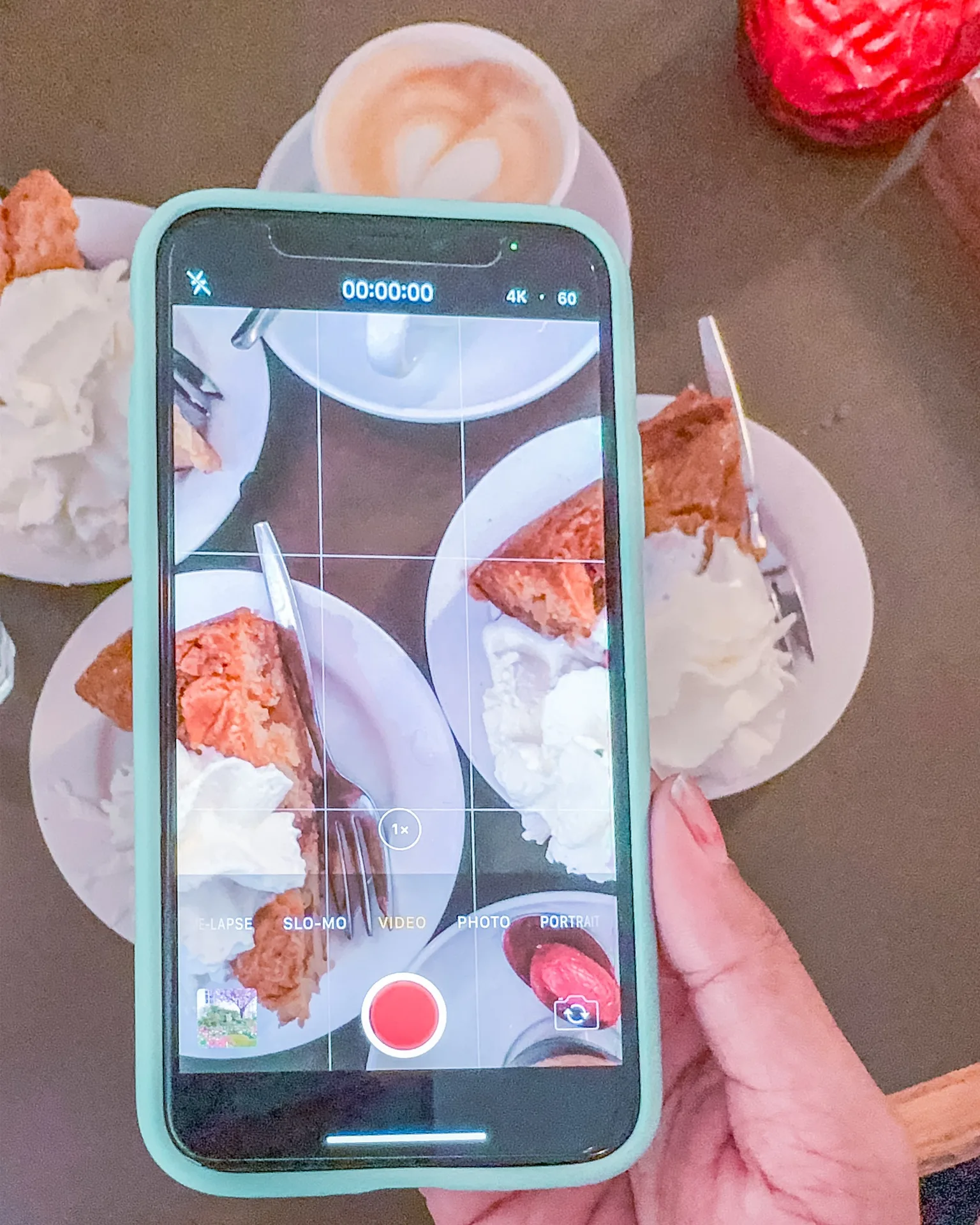 Hand holding a phone filming plates of cake with whipped cream and a latte on a café table.