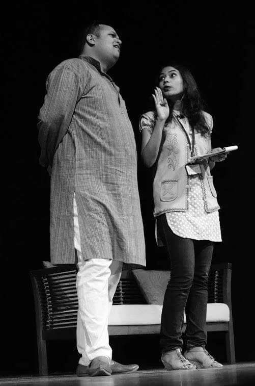 Two people in traditional and casual attire stand on a stage, the woman animatedly speaking while holding a notebook beside a bench.