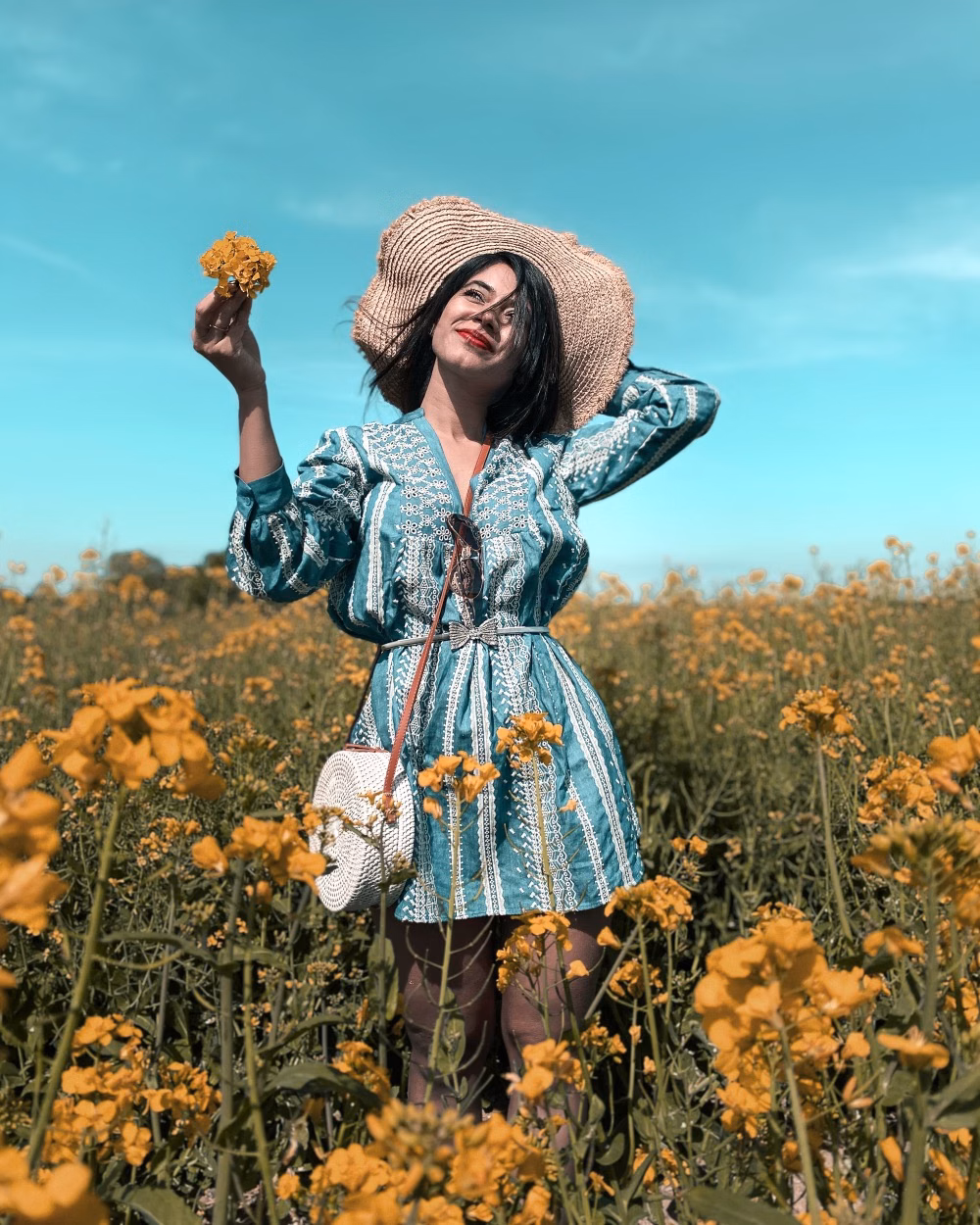 Woman in a straw hat and blue dress standing in a field of yellow flowers under a clear sky