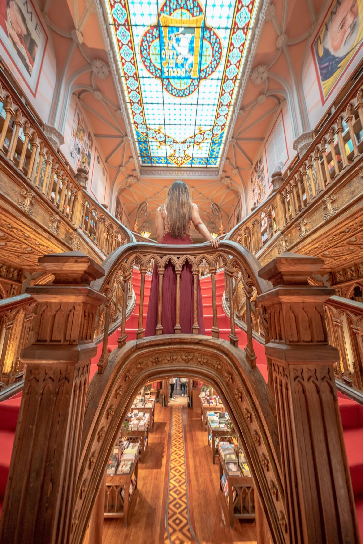 Visitor in a red dress stands on an ornate wooden staircase beneath a stained-glass skylight inside a grand historic bookstore.