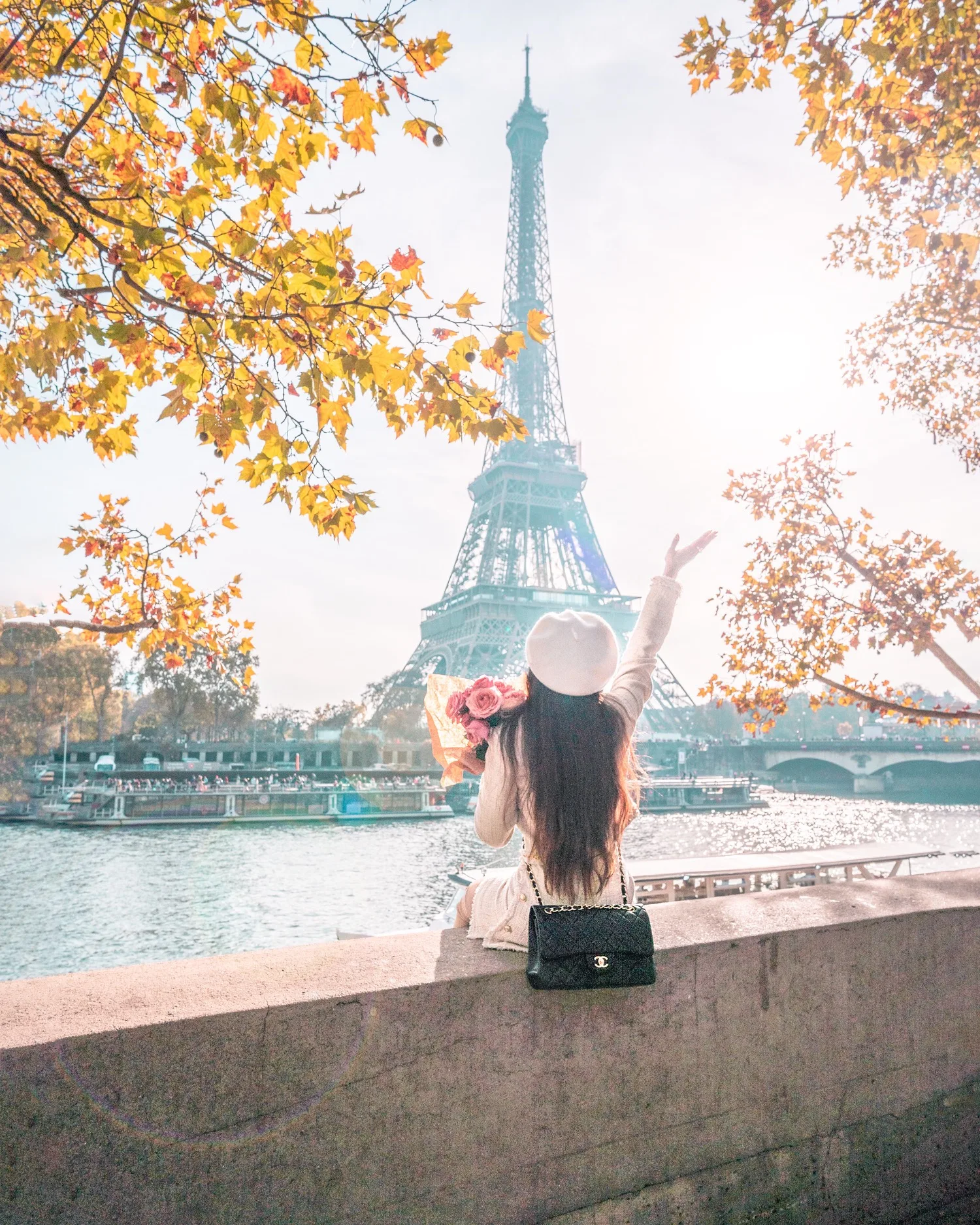 Woman in a beret holding flowers and waving toward the Eiffel Tower across the Seine framed by autumn leaves