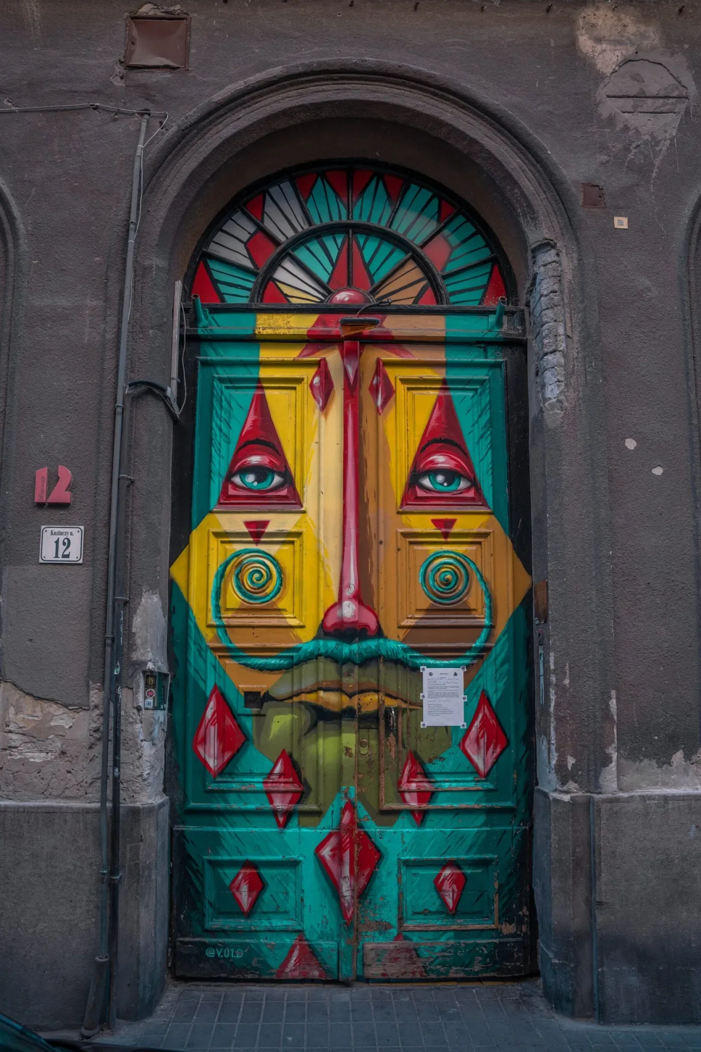 Colourful street-art portrait painted across an ornate wooden doorway, featuring a stylised face with teal mustache, spiral cheeks and geometric red accents.