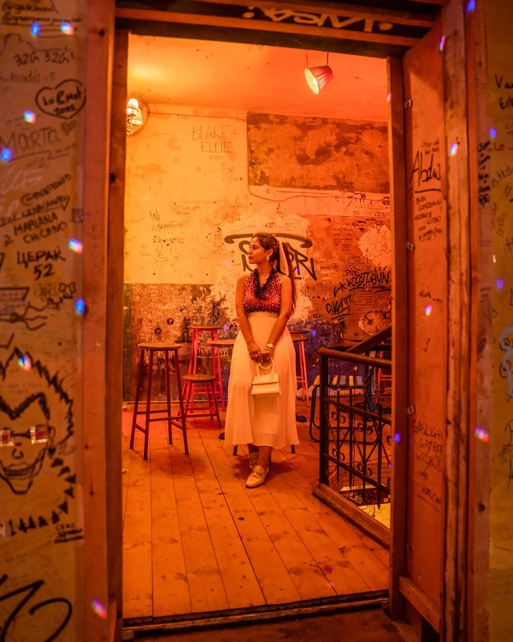 Woman in a white skirt and patterned top holding a small bag, standing in a graffiti-covered, warmly lit bar room.