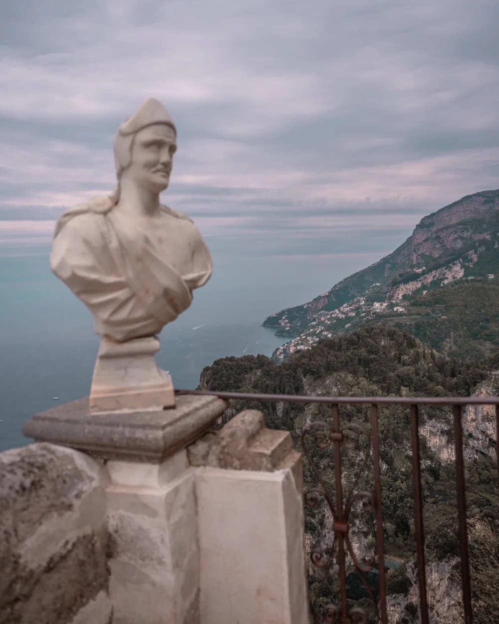 Marble bust on a terrace overlooking dramatic coastal cliffs and the sea under a cloudy sky.