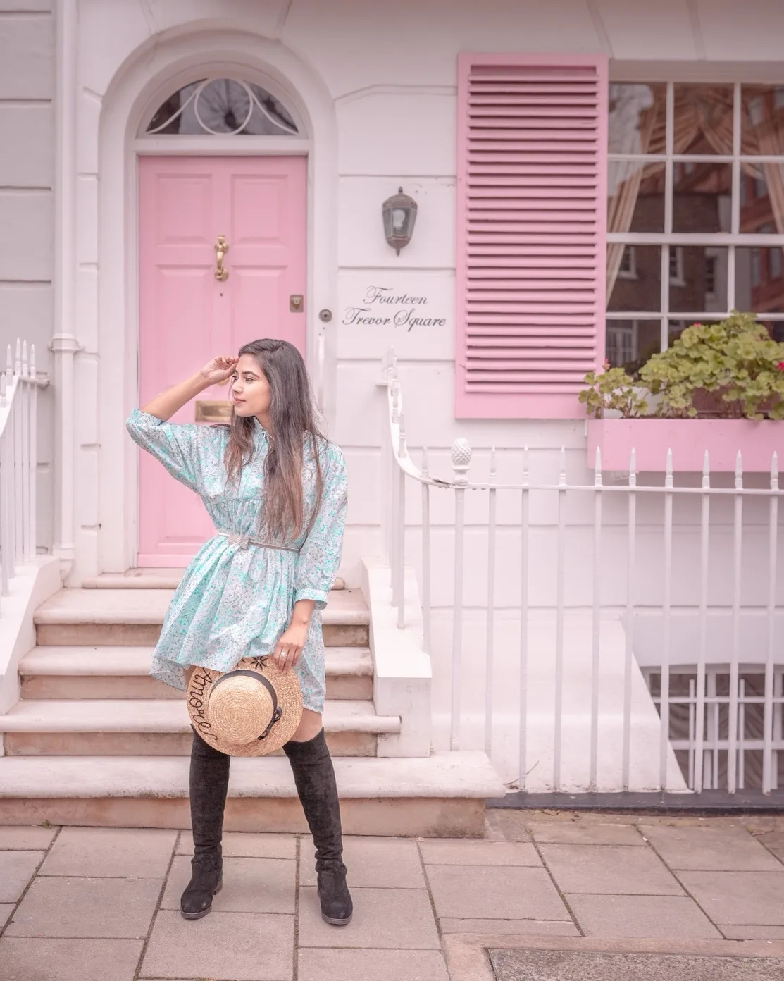Young woman in a floral dress and knee-high boots holding a straw hat, posing on steps before a pastel pink door and shuttered window.