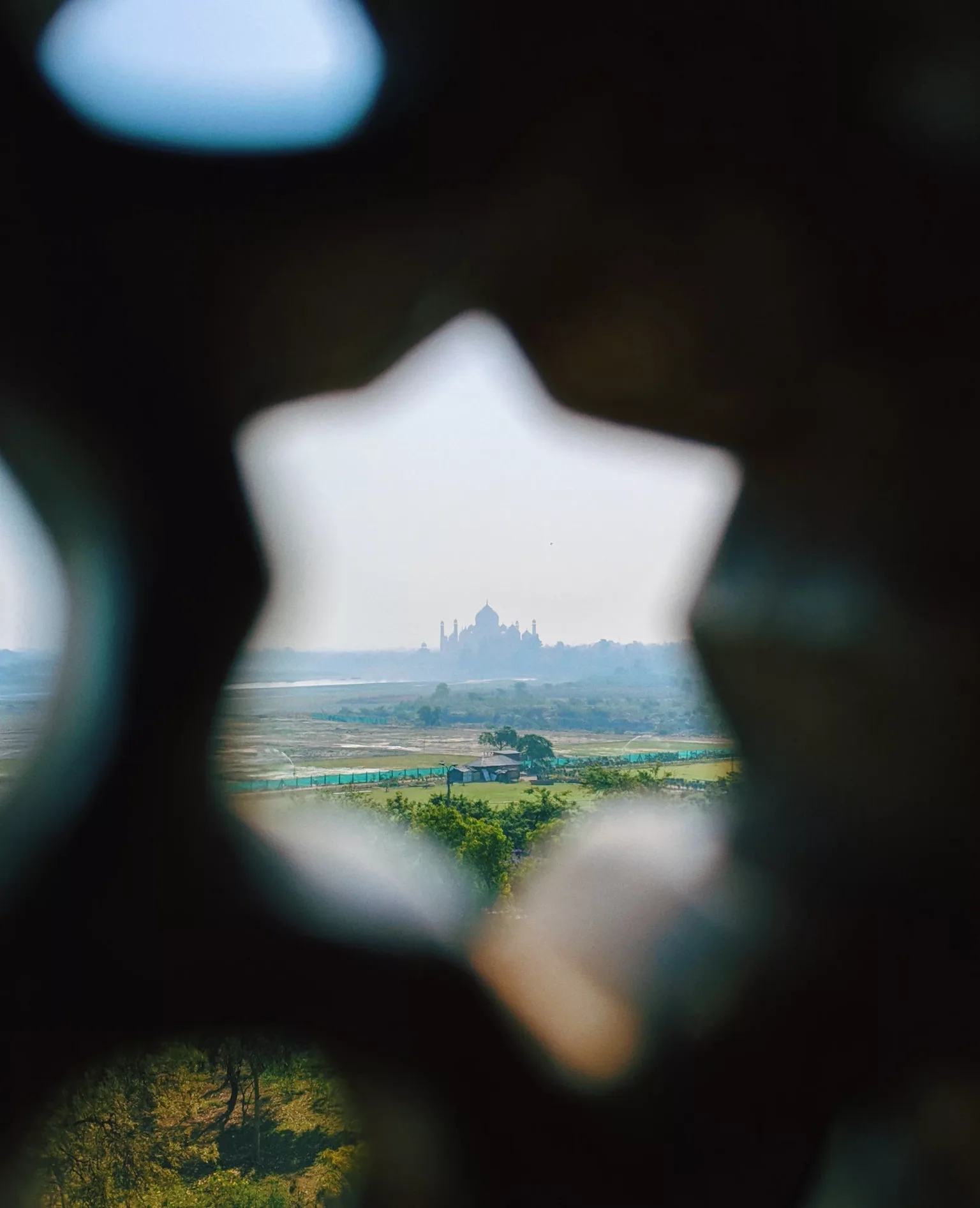 Distant white mausoleum silhouette framed through a star-shaped lattice overlooking green fields and a hazy river plain