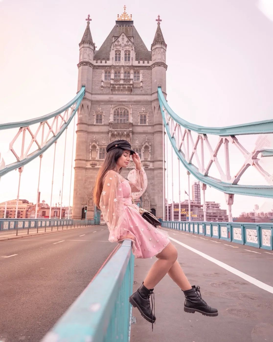 Tower Bridge at sunrise in London ; Young woman in a pink dress and black cap perched on the railing of Tower Bridge with the bridge's Gothic towers and teal suspension cables behind her.
