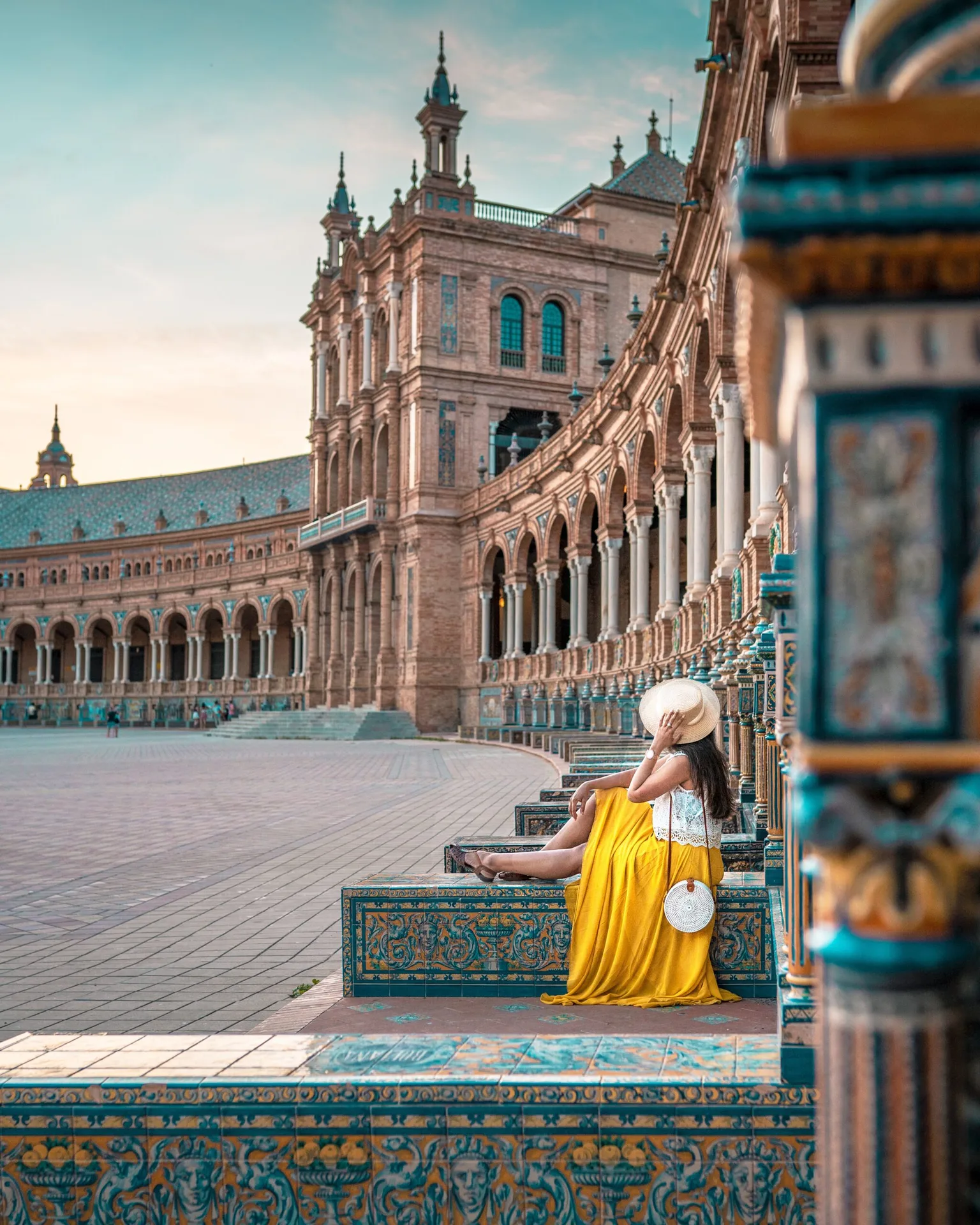 Woman in a yellow skirt and sunhat sitting on decorative tiled benches under the arched colonnade of Plaza de España, Seville.