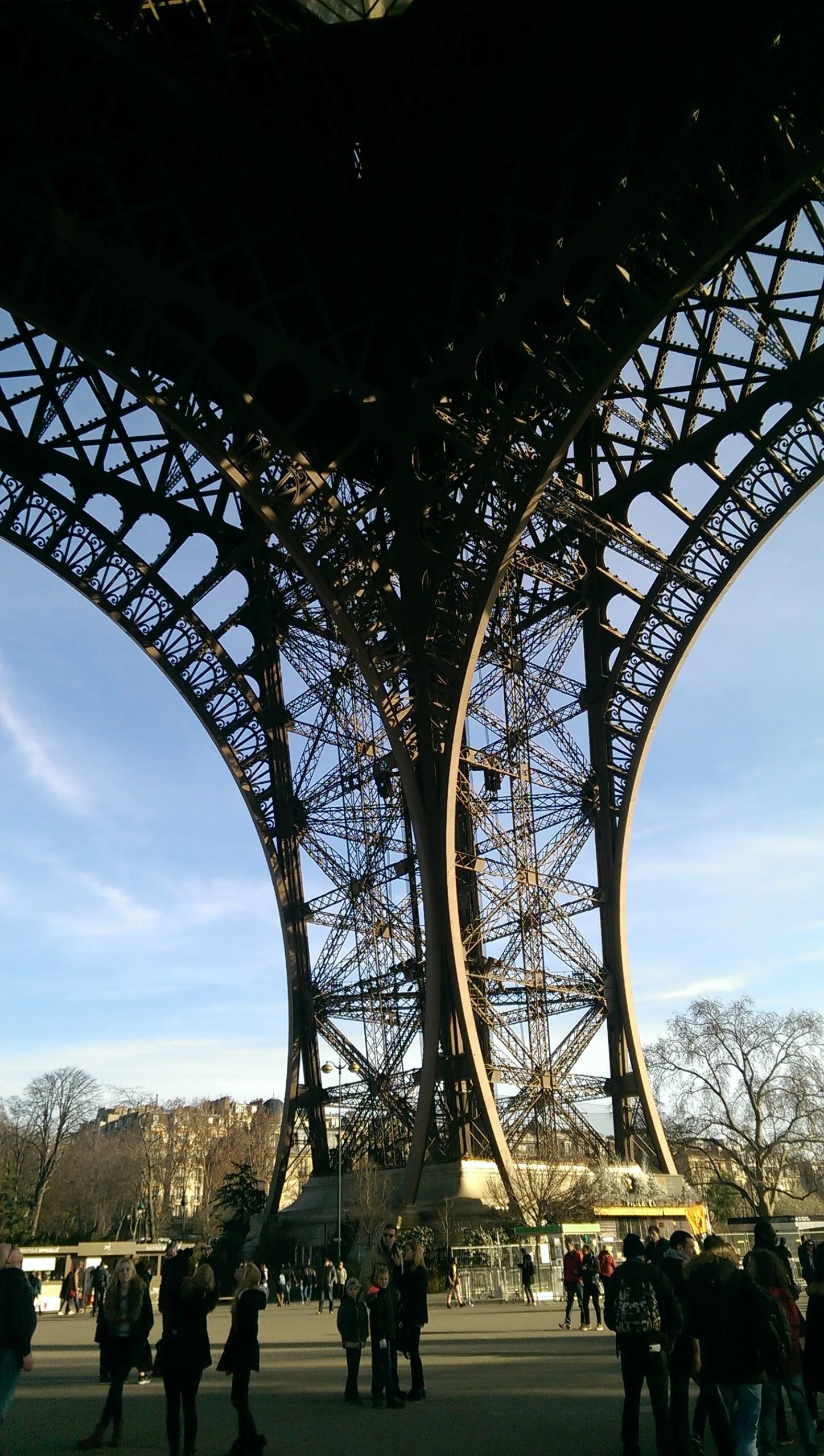 View upward through the ornate iron arches and latticework of the Eiffel Tower with visitors gathered beneath on a clear day.