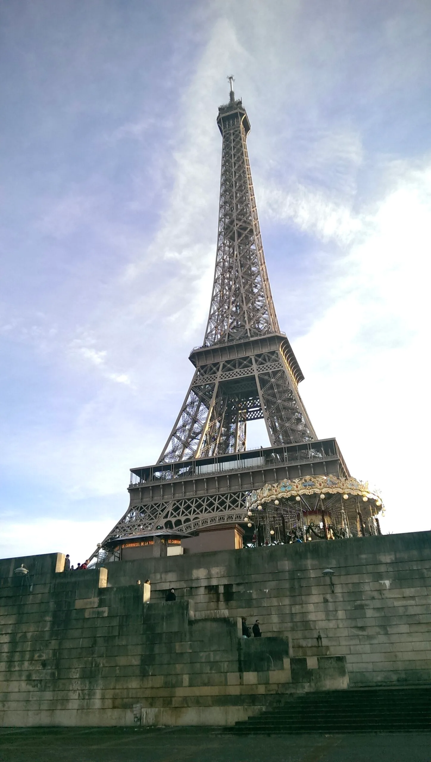Eiffel Tower rising above stone terraces with a small ornate carousel at its base under a pale blue sky.