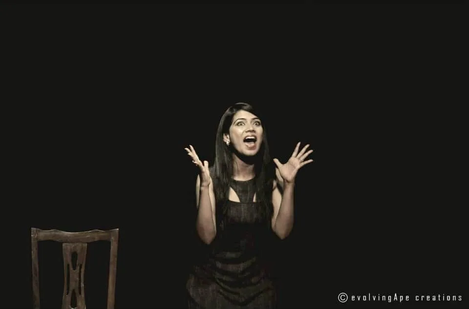 Woman on a dark stage with animated expression and raised hands, seated beside an empty wooden chair.