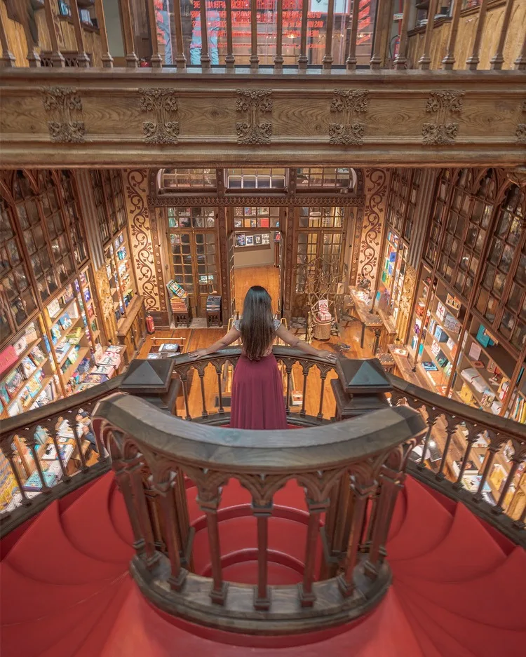 Woman in a red dress standing on an ornate wooden balcony overlooking a grand, multi-level historic bookstore filled with books.