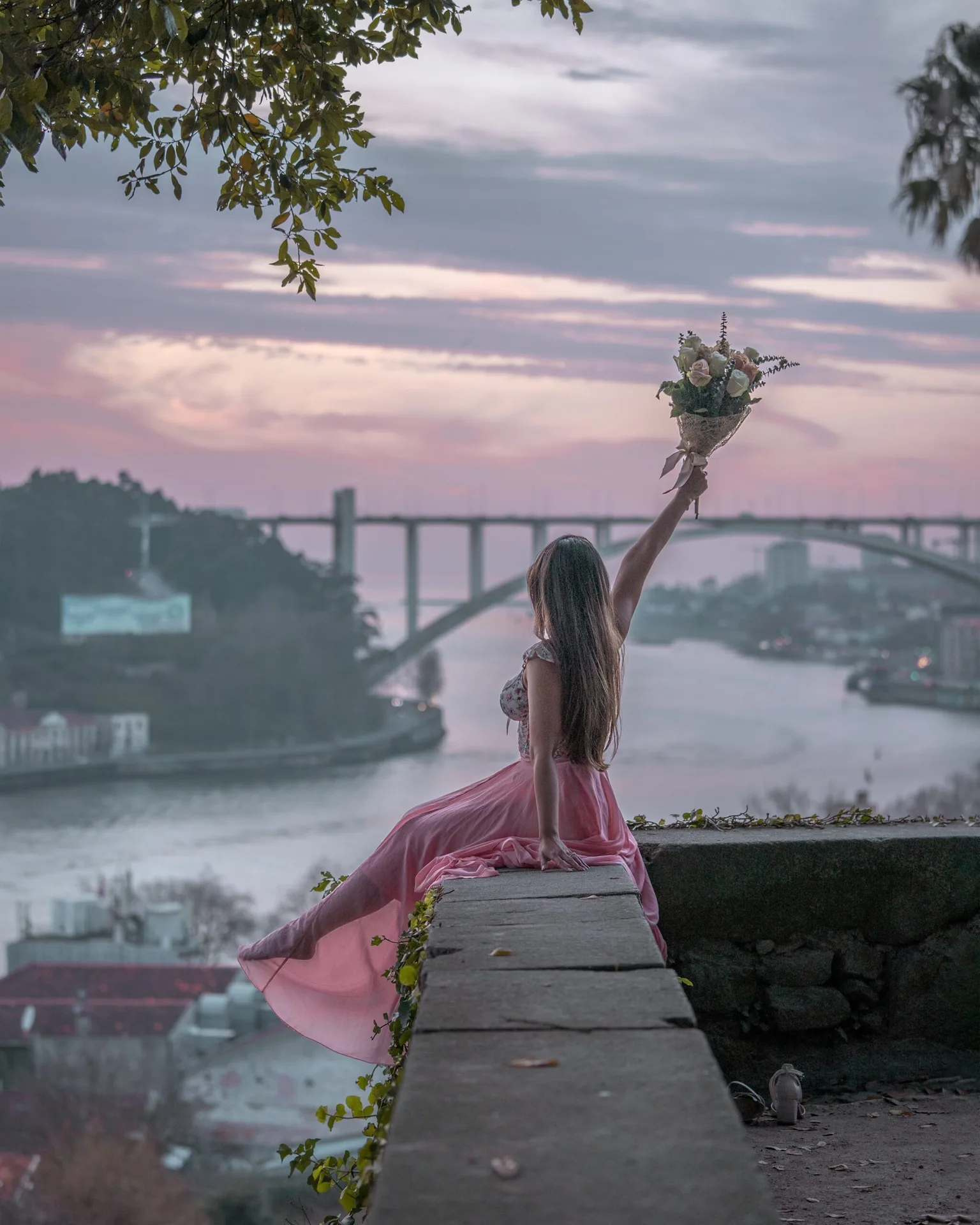 Woman in a flowing pink dress sits on a stone ledge overlooking a river and distant bridge at sunset, holding a bouquet aloft.