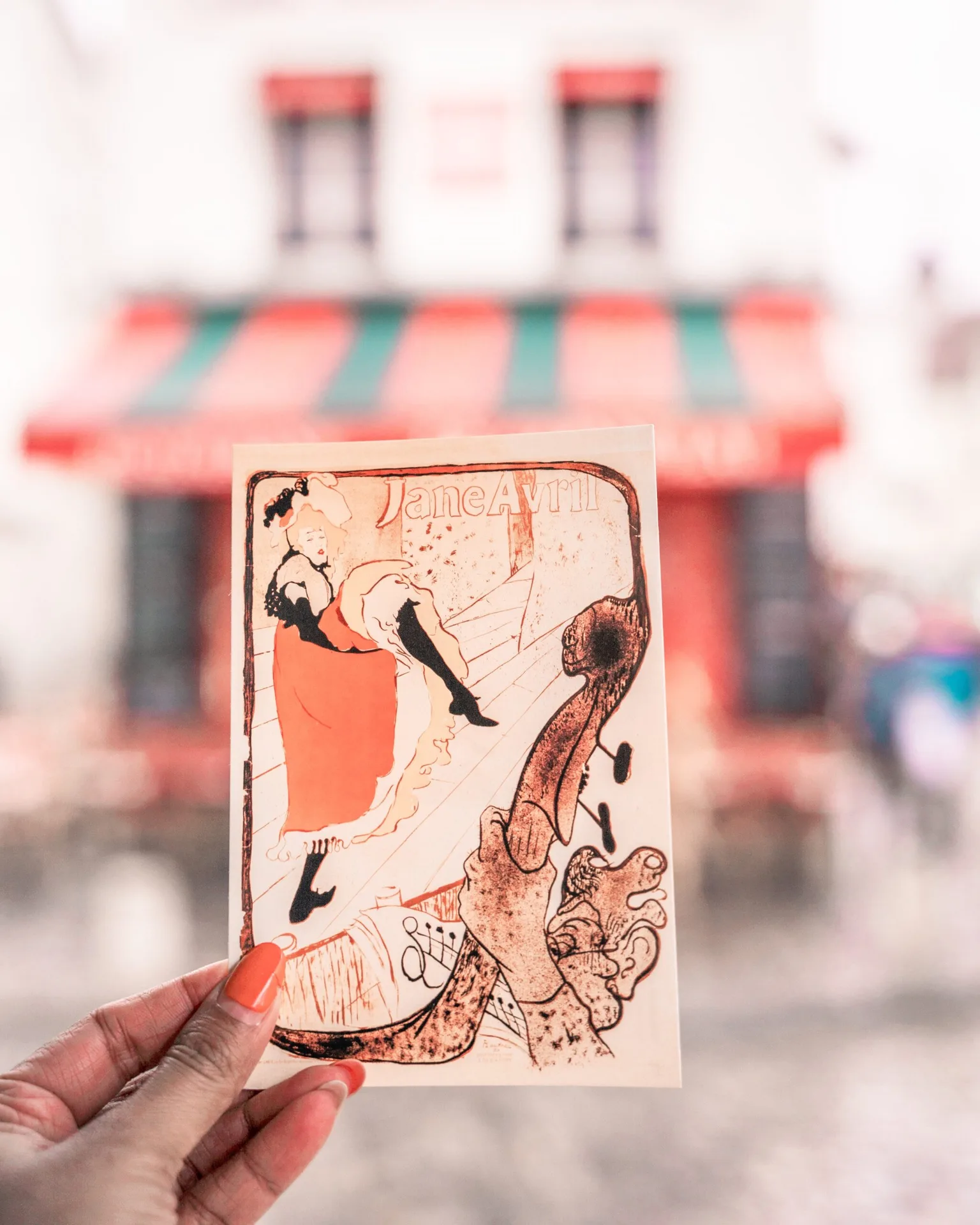 Hand holding a vintage-style Jane Avril postcard with a blurred café awning in the colourful background.