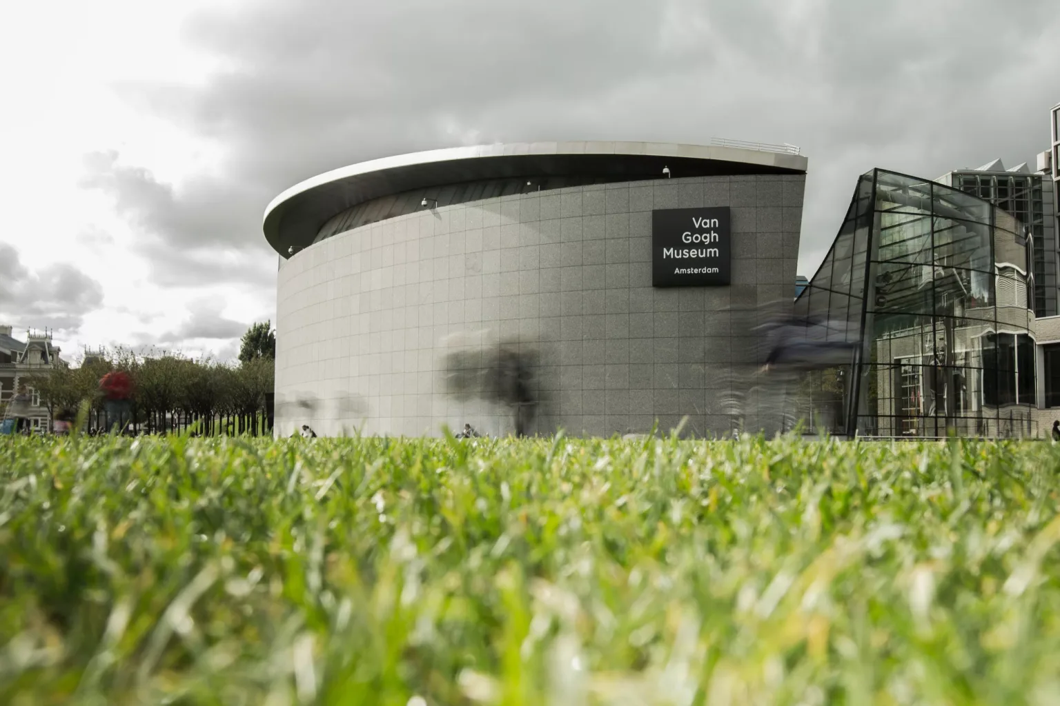 Low-angle view of the circular Van Gogh Museum building and glass entrance with blurred visitors on a cloudy day.