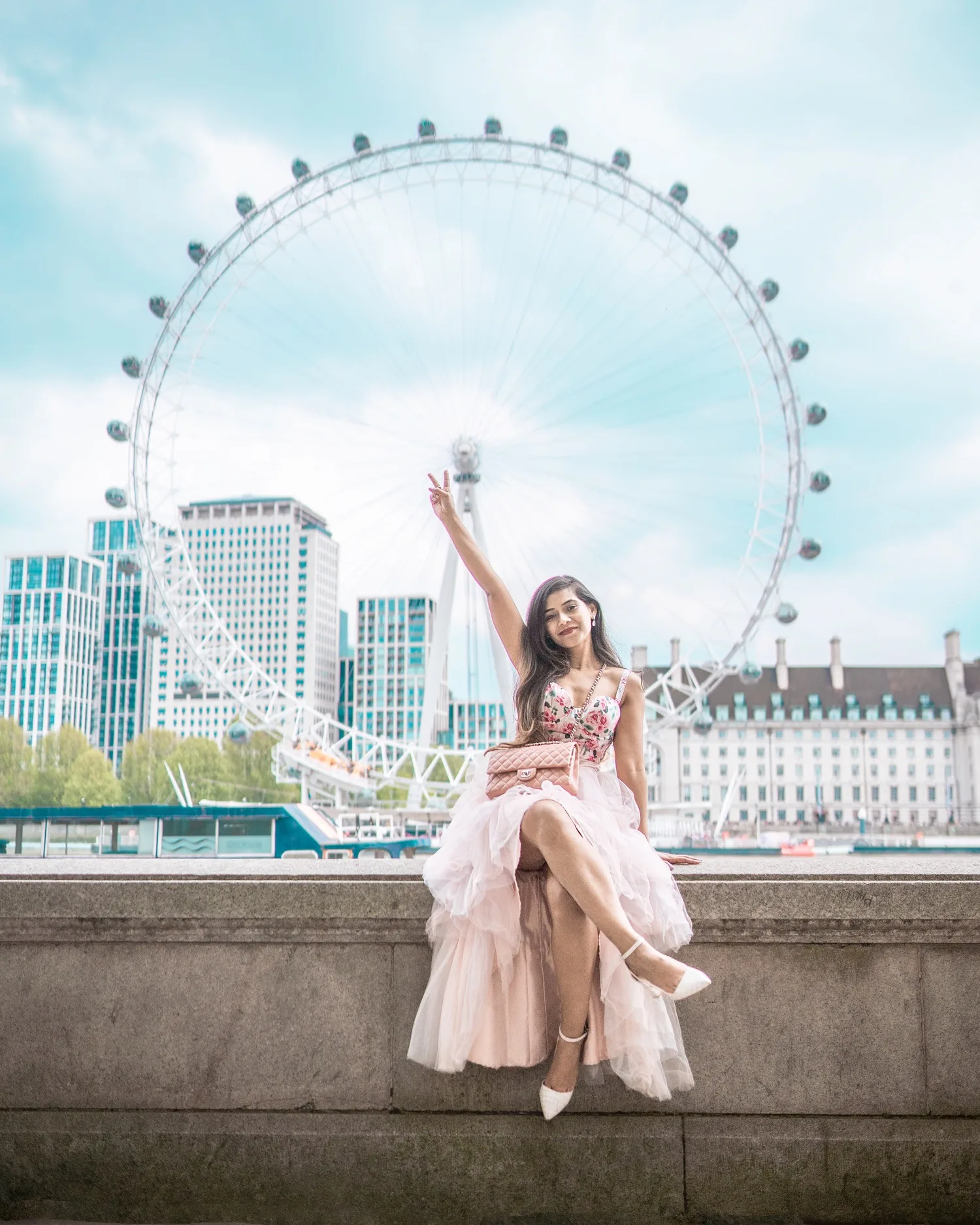 Useful travel Links - Young woman in a pink tulle dress sitting on a riverside wall with the London Eye ferris wheel rising behind her as she flashes a peace sign.