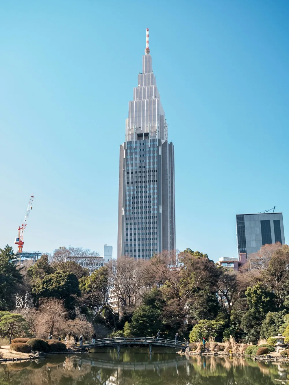 Tall modern skyscraper rising behind a tranquil Shinjuku Gyoen park pond and wooden footbridge under a clear blue sky in Tokyo.