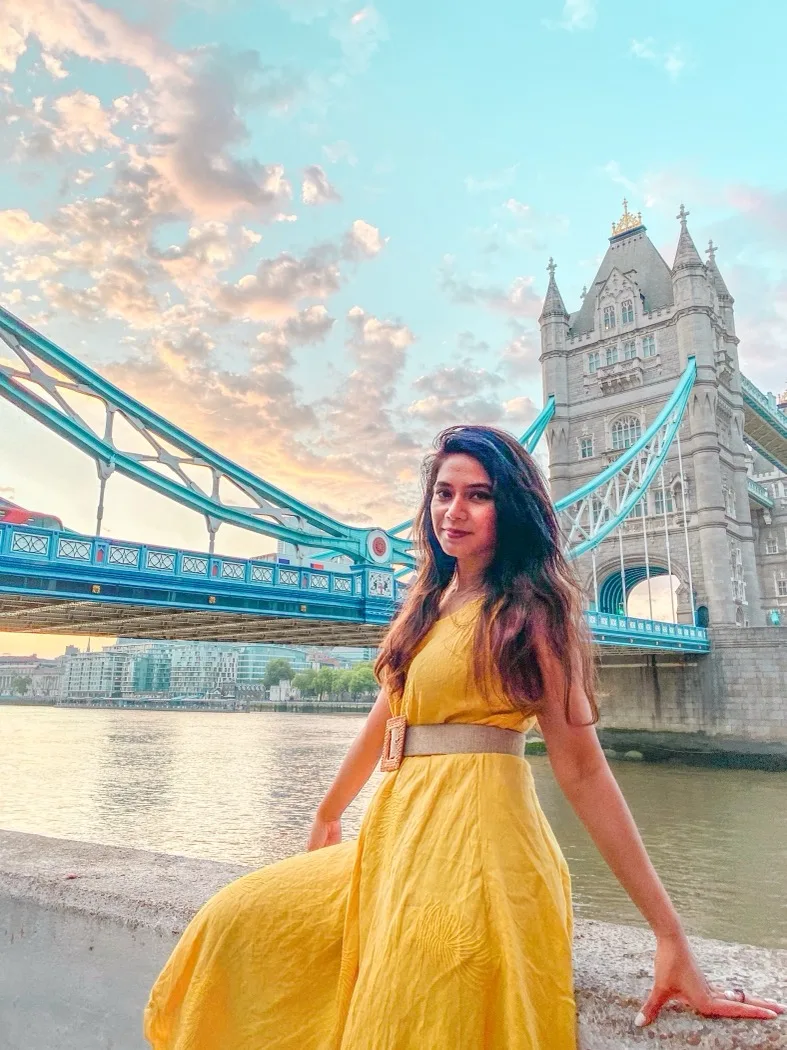 Woman in a yellow dress posing by the River Thames with Tower Bridge and a pastel sunset sky in the background.