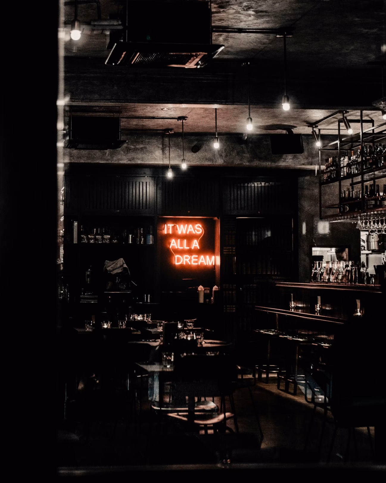 Dim bar interior with hanging lights and a neon sign reading It Was All a Dream