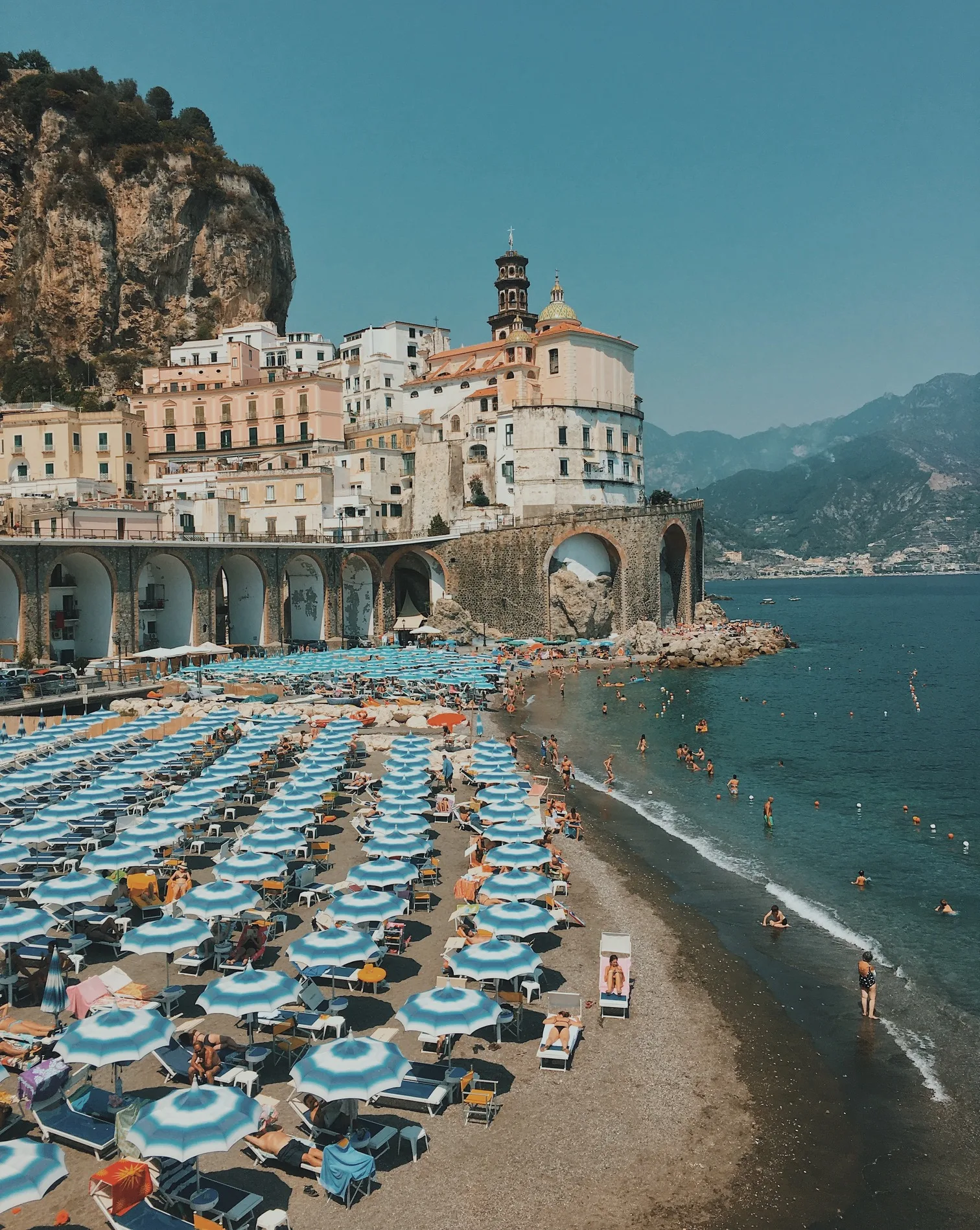 Sun-drenched Amalfi Coast beach with rows of blue-and-white umbrellas, swimmers in the turquoise sea, and cliffside Mediterranean buildings.