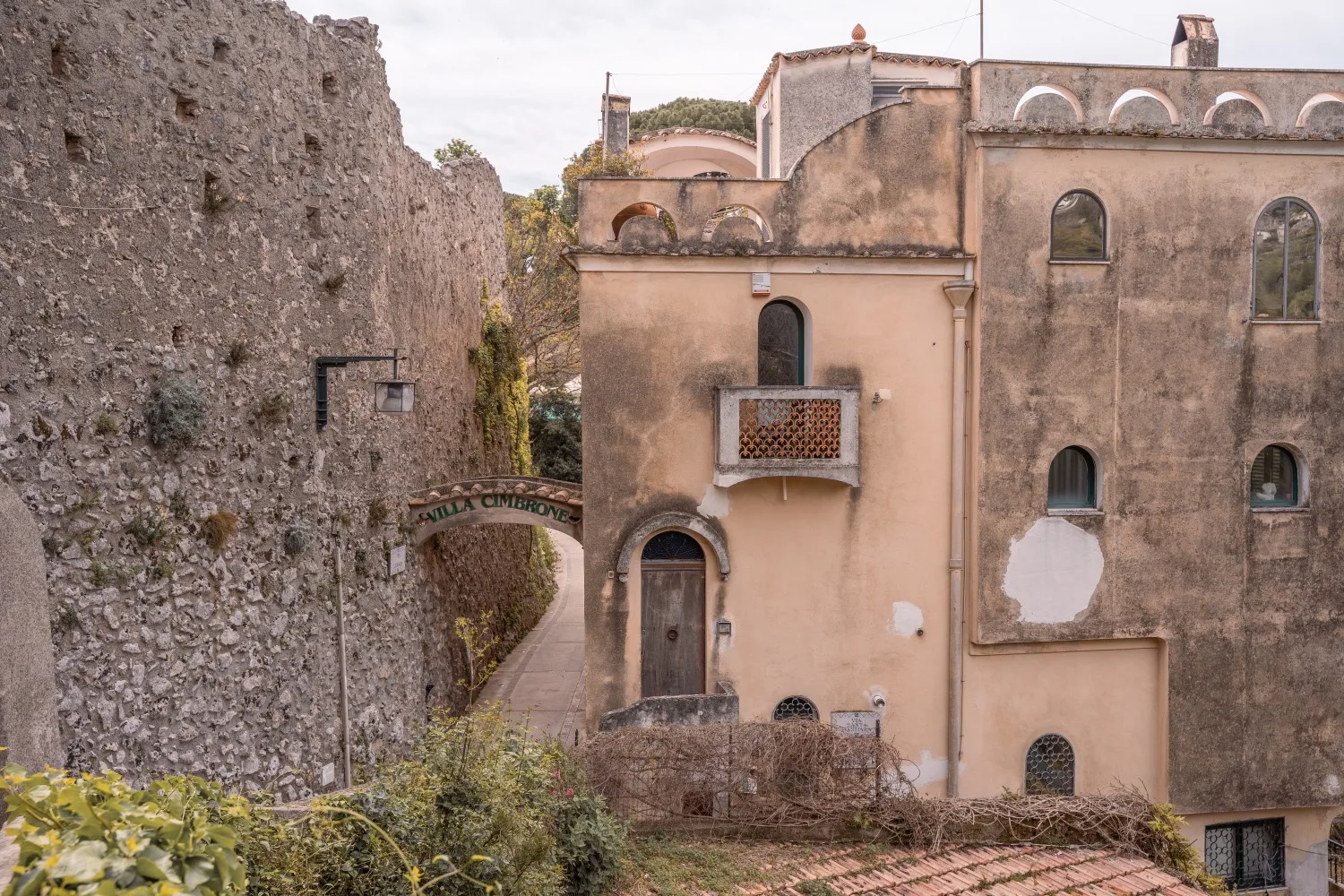 Weathered ochre villa facade with arched windows and small balcony beside an old stone wall and narrow shaded alley marked "Villa Cimbrone."
