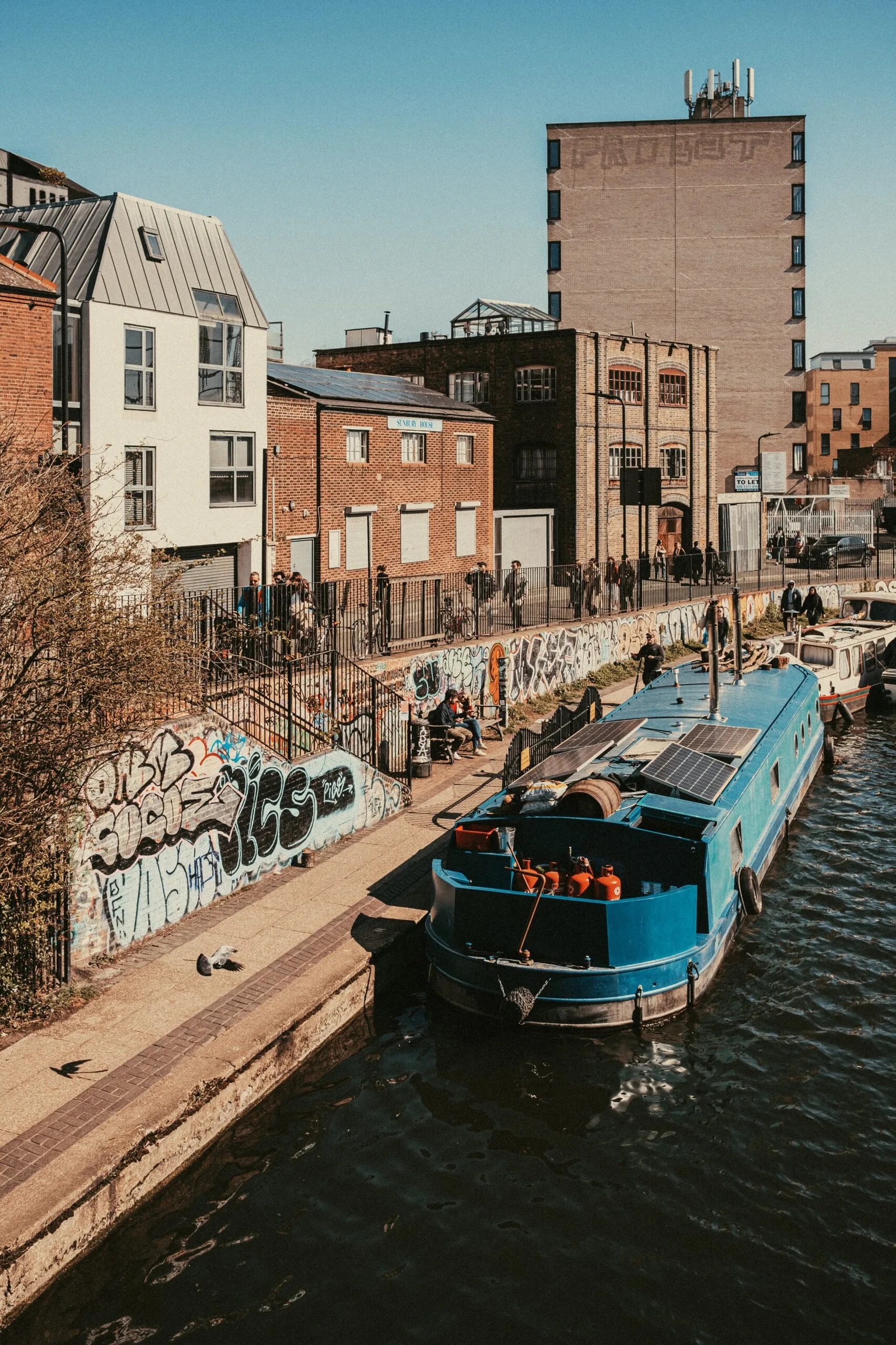 Narrow blue canal boat moored beside a graffiti‑covered towpath with brick warehouses and pedestrians in the background.