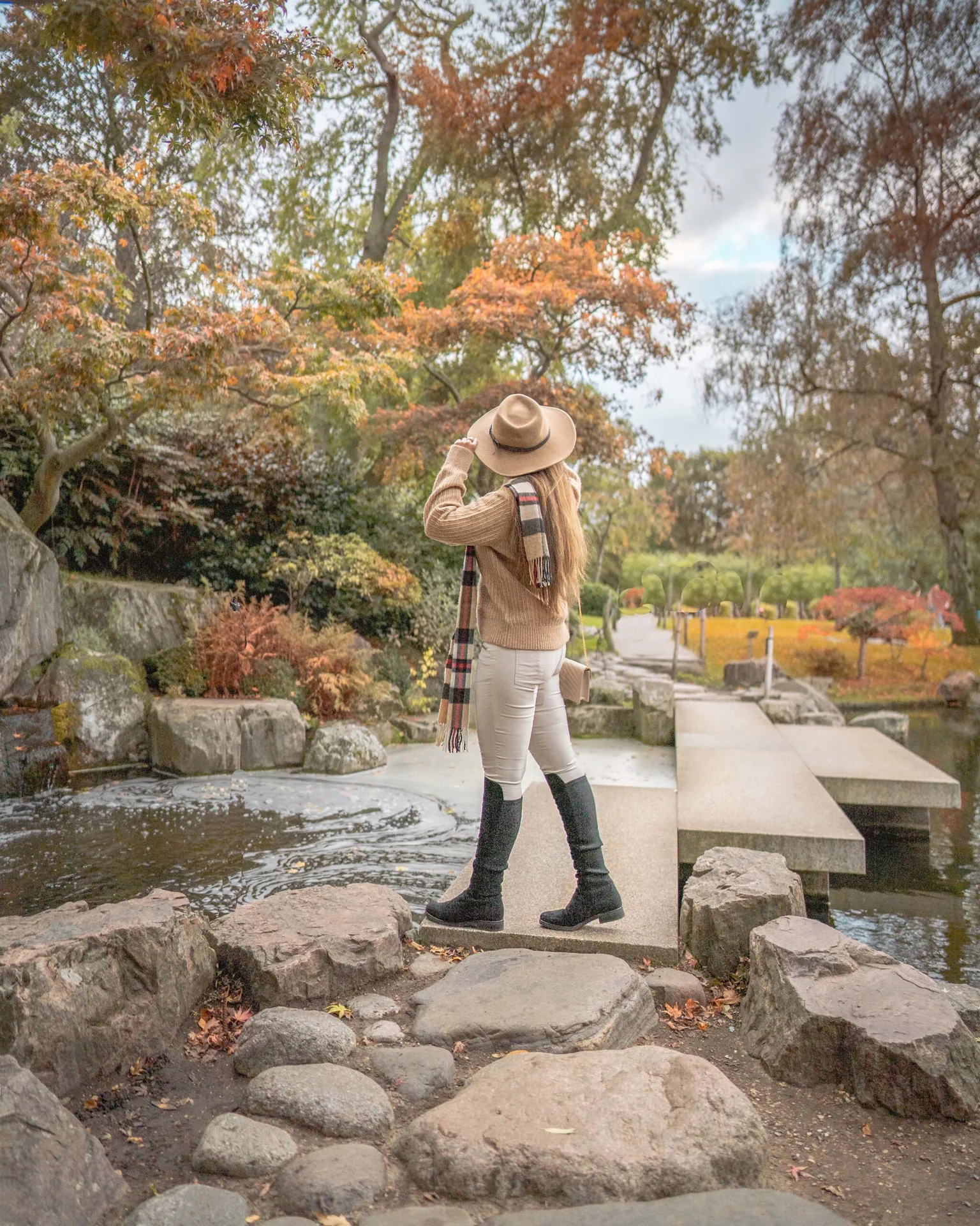 Woman in a hat and scarf standing on a stone bridge over a pond, surrounded by autumn trees and gardens.