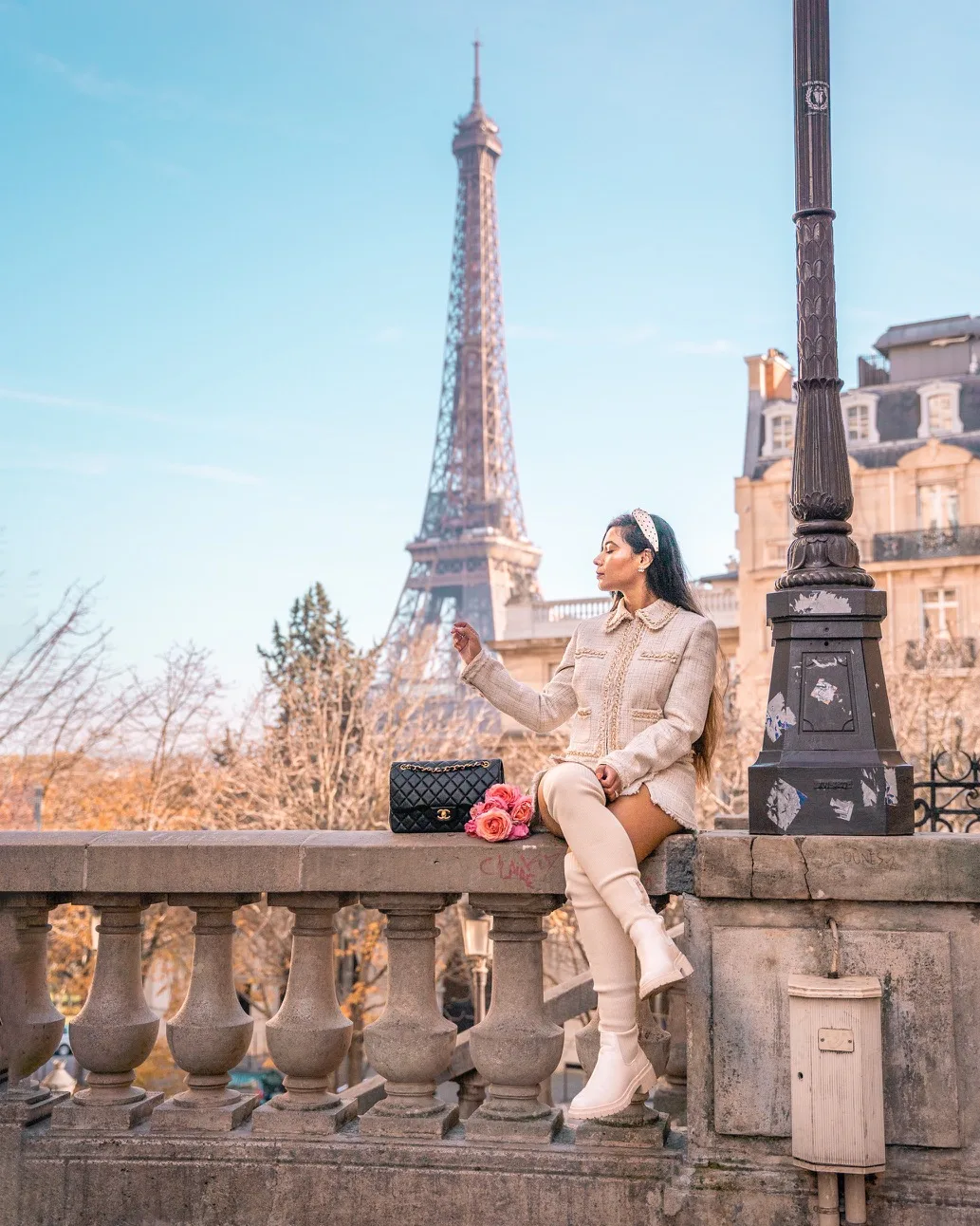 Woman in cream coat and knee-high boots sitting on a Paris balustrade with a bouquet and Chanel bag, Eiffel Tower rising in the background.