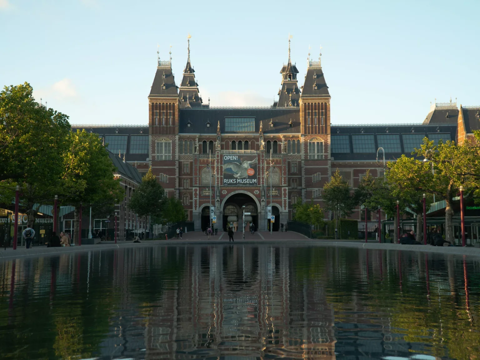 Grand Rijksmuseum building with ornate towers reflected in a calm rectangular pond in front, framed by trees.
