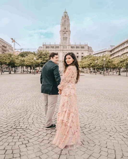 Couple holding hands and looking back toward the camera in a spacious cobblestone plaza with a grand clock tower and trees in the background.