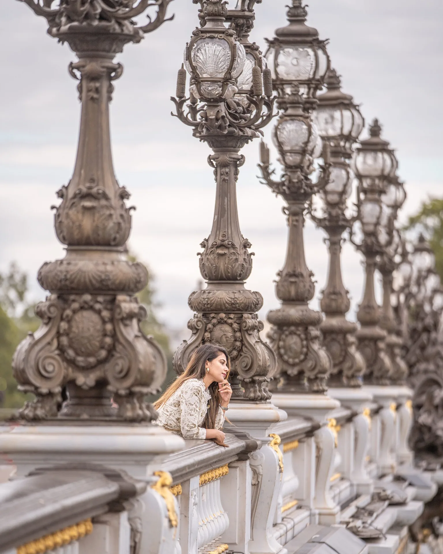 Woman leaning on ornate balustrade beneath a row of decorative bronze lamp posts on an elegant European bridge