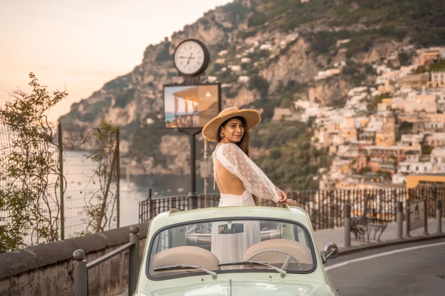 Affordable Luxury Travel in Positano, Amalfi Coast, Woman in a straw hat and backless white dress leaning from a mint-green vintage convertible overlooking a coastal cliffside town at sunset