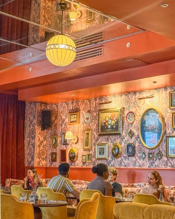 Guests chatting in a cosy vintage café with patterned wallpaper, framed art and warm hanging lights