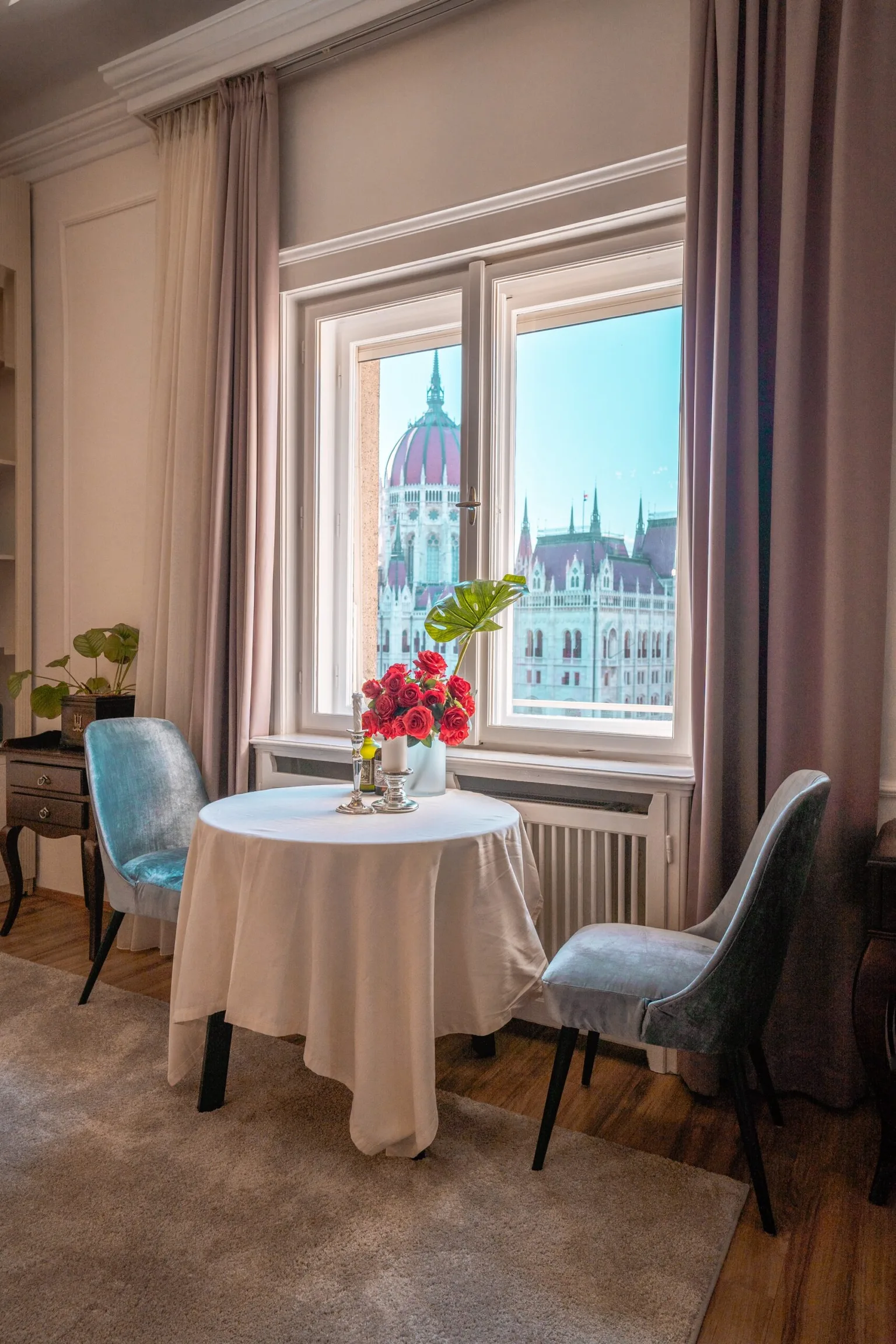 Small dining table with a white cloth, vase of red roses and two velvet chairs by a window overlooking a domed historic building.