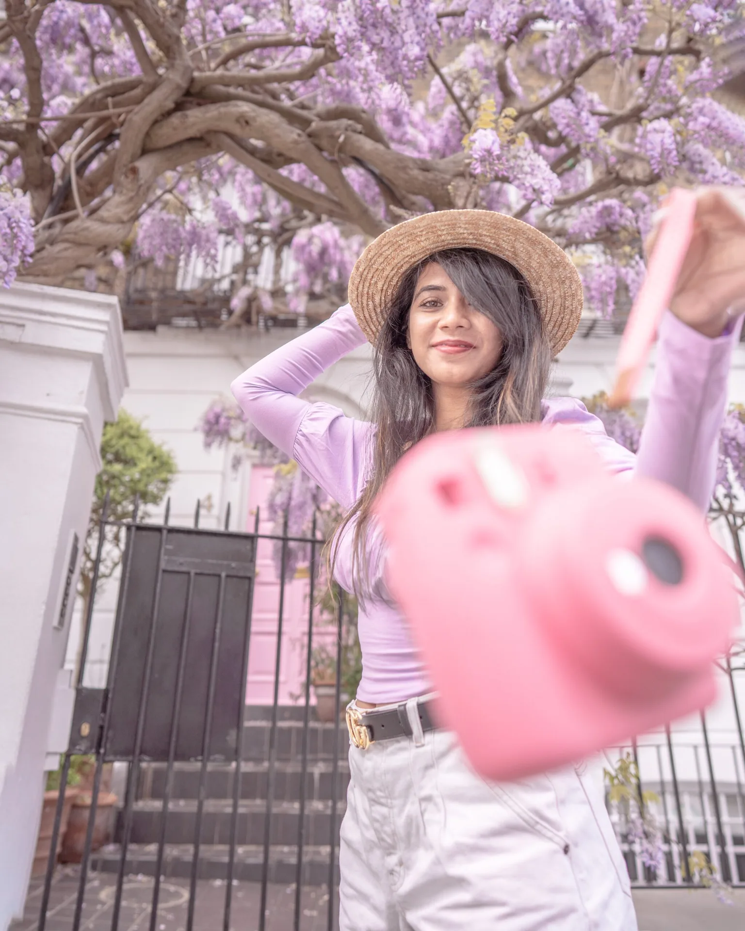 Woman in a straw hat smiling and holding out a pink instant camera under blooming wisteria in front of a pastel townhouse.