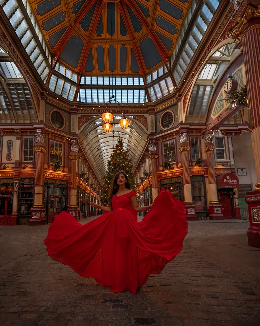 Woman in a flowing red gown standing beneath a decorated glass-roofed Victorian arcade with a large Christmas tree and warm hanging lamps