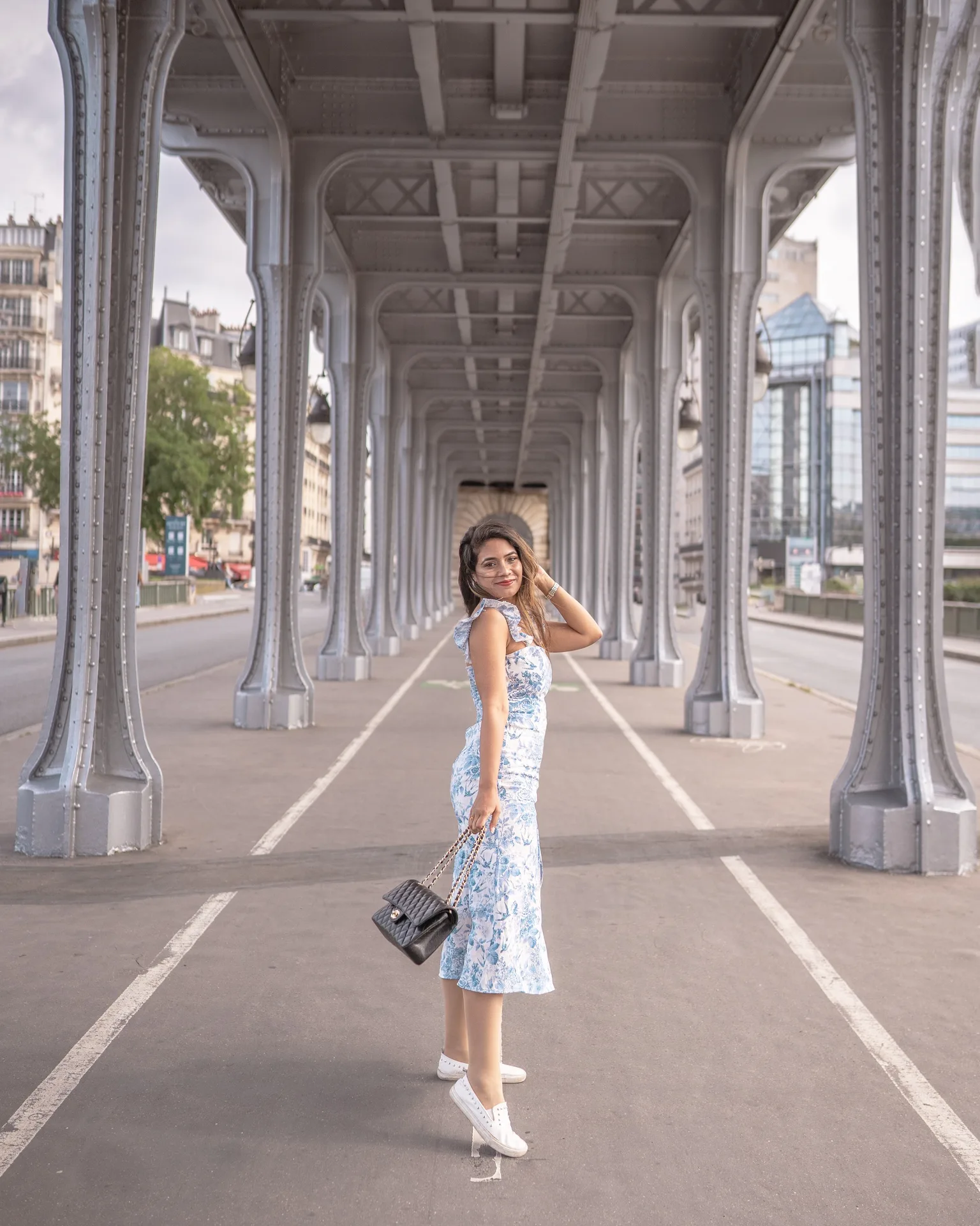 Woman in a blue floral midi dress and white sneakers holding a black quilted handbag while posing under the iron arches of a Parisian bridge.
