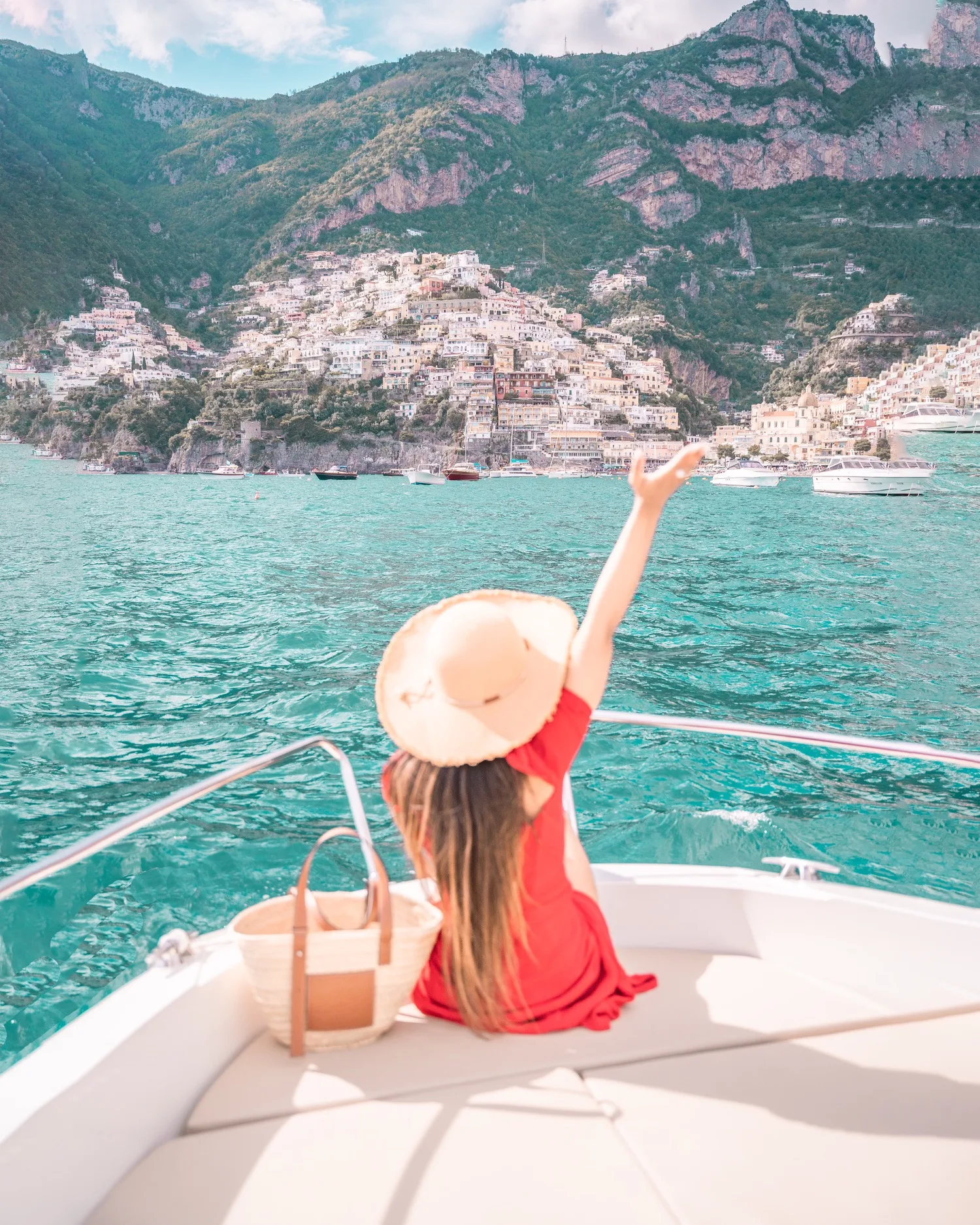 Woman in a red dress and straw hat sitting on a boat, waving toward pastel cliffside buildings along a turquoise coastline.