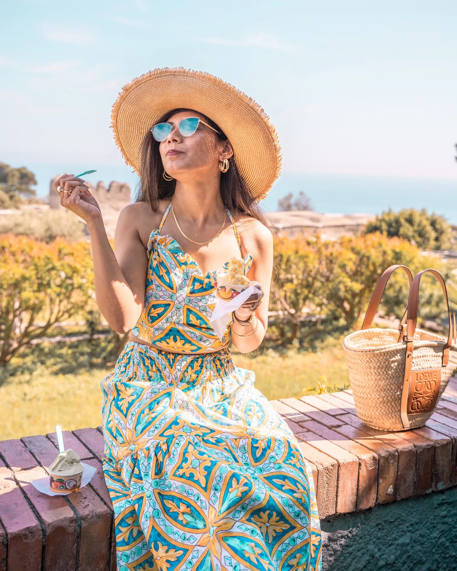 Woman in a patterned summer dress and wide straw hat enjoying gelato on a sunny coastal hill with a woven tote beside her