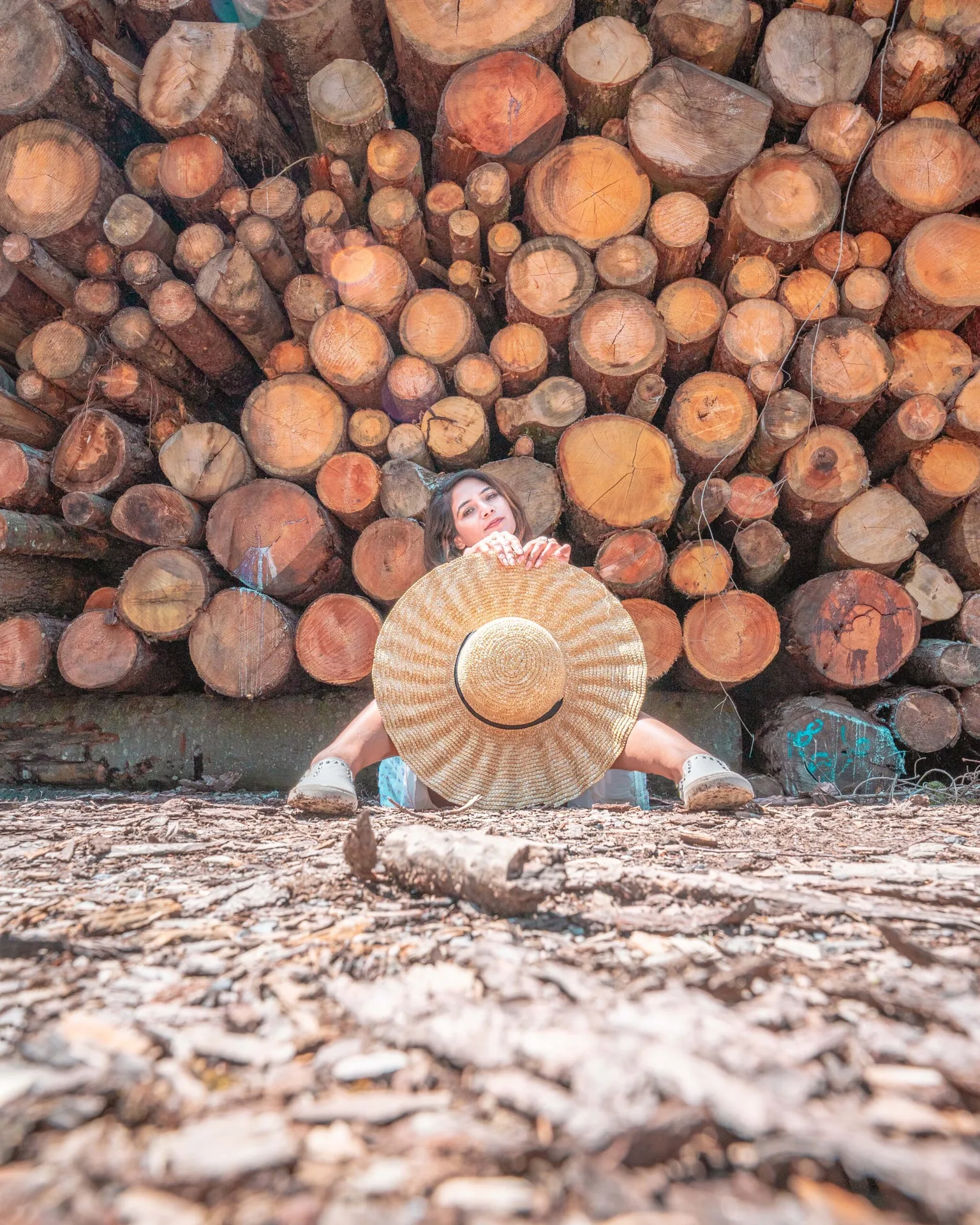 Woman sitting in front of a large stack of cut logs holding a wide straw sunhat that partly hides her face.