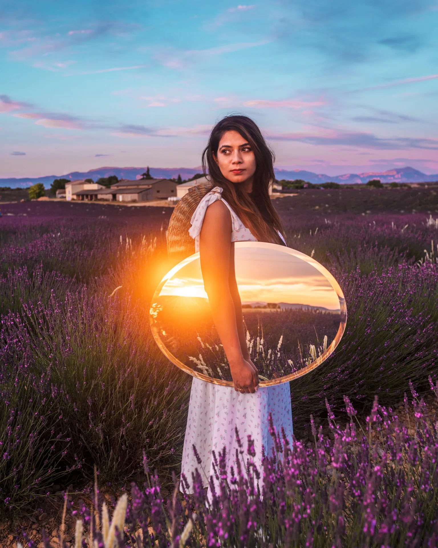 Young woman in a white sundress holding a reflective oval mirror in a lavender field at sunset, the sky and sun glowing in the mirror.