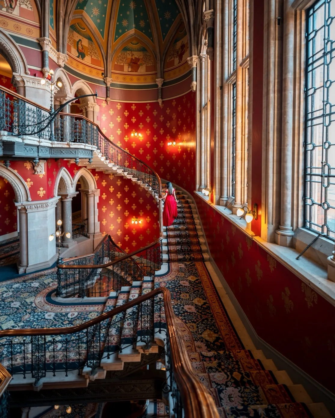 Woman in a flowing red dress ascending an ornate curved staircase in a grand, richly decorated historic interior with tall leaded windows.