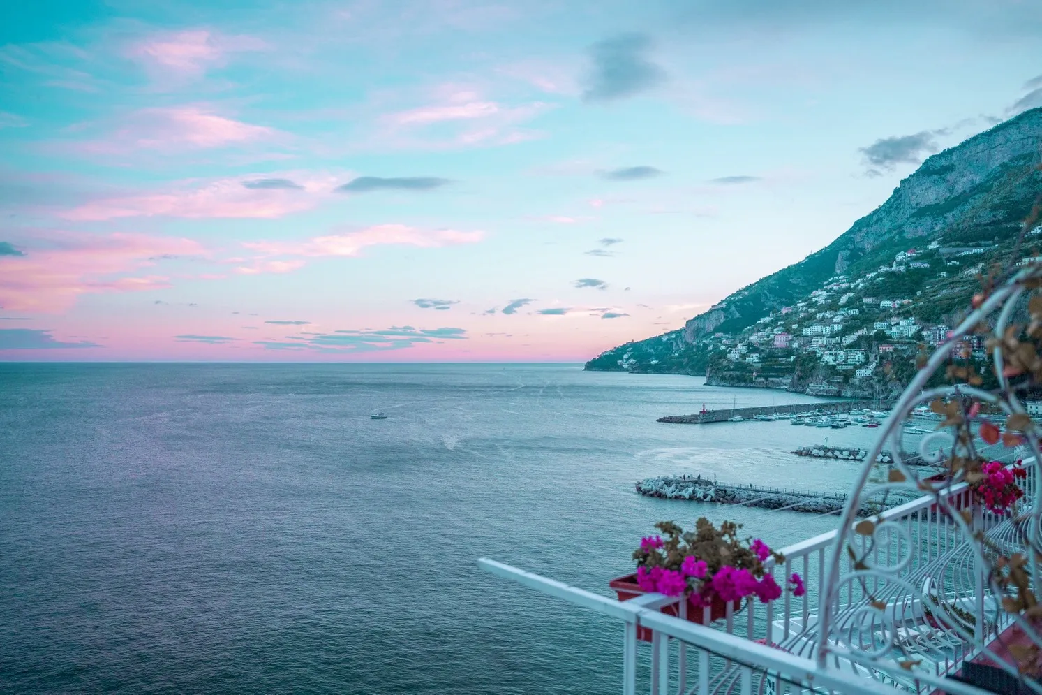 Seaside view from a balcony with pink flowers overlooking a calm bay, coastal cliffs and pastel sunset sky.