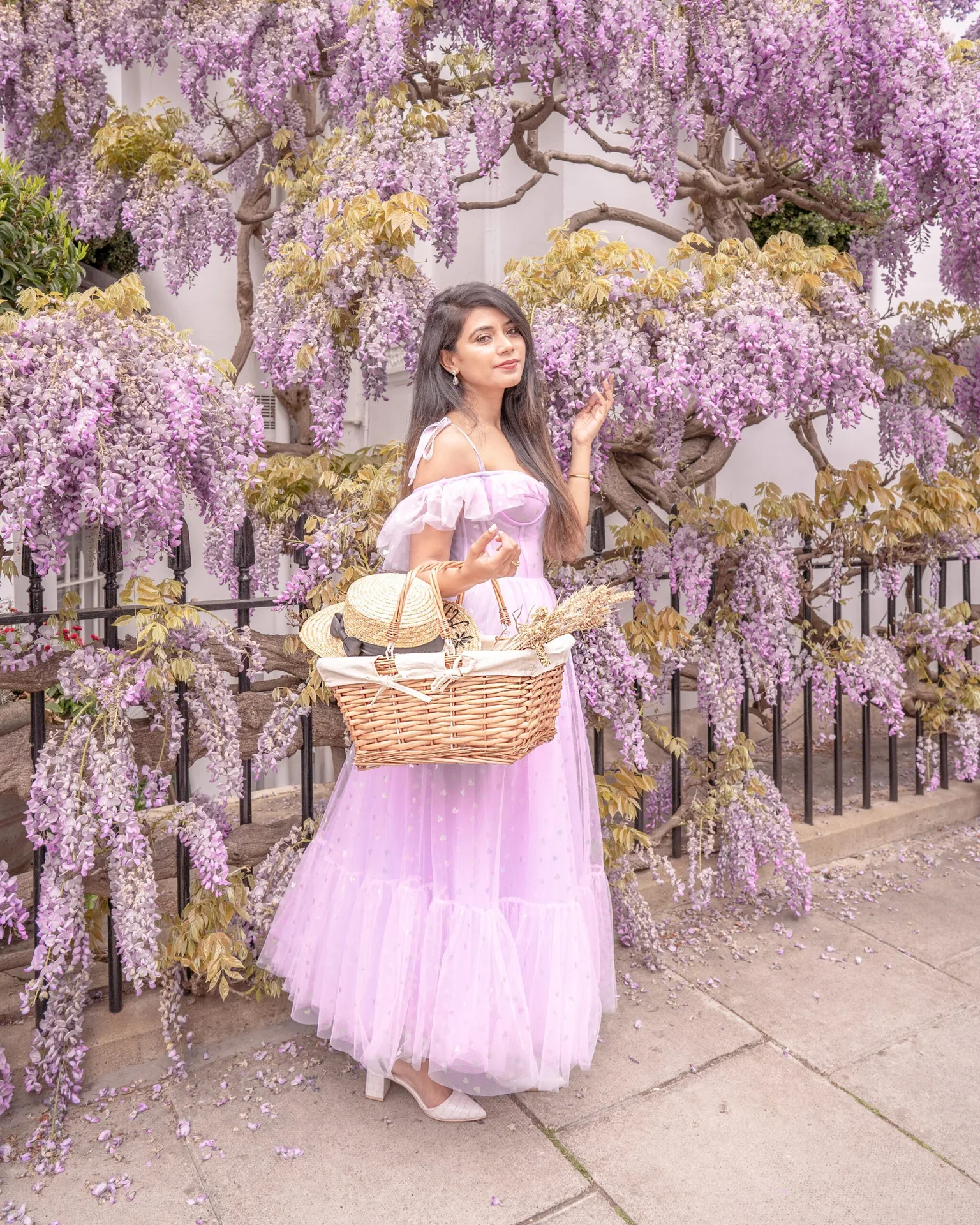 Woman in a flowing lilac dress holding a wicker picnic basket beneath cascading wisteria blossoms along a city pavement