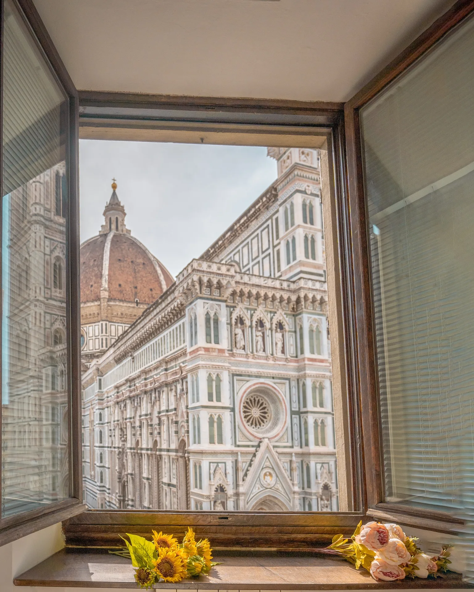View of Florence's Cathedral of Santa Maria del Fiore dome and ornate façade seen through an open window with sunflowers and roses on the sill.